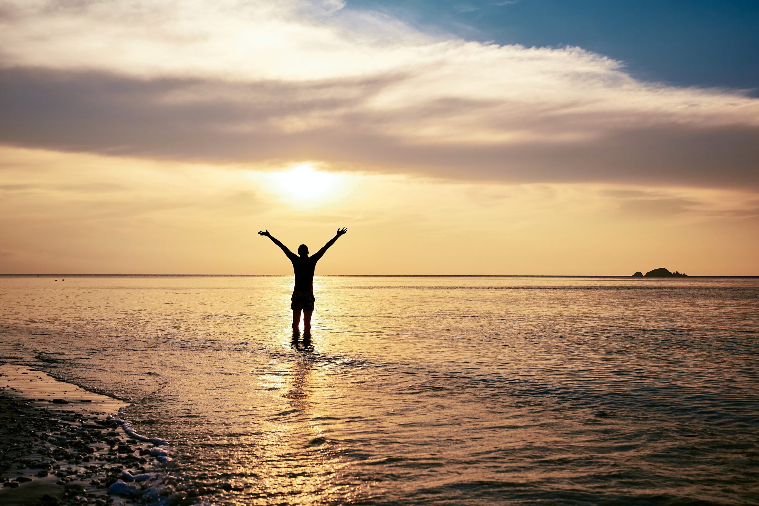 a rear view of a happy man with arms outstretched standing in the sea during sunset, as part of whole person therapy